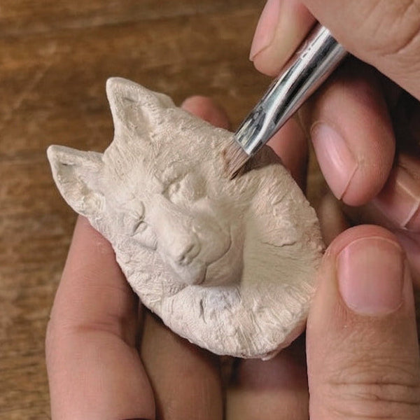 A close-up of a person's hands carving a small white ceramic or clay Husky head figurine using a fine metal tool over a rustic wooden surface.